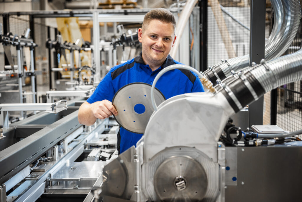 Techniker bedient eine Maschine mit einem großen runden Metallbauteil in einer industriellen Produktionsanlage.